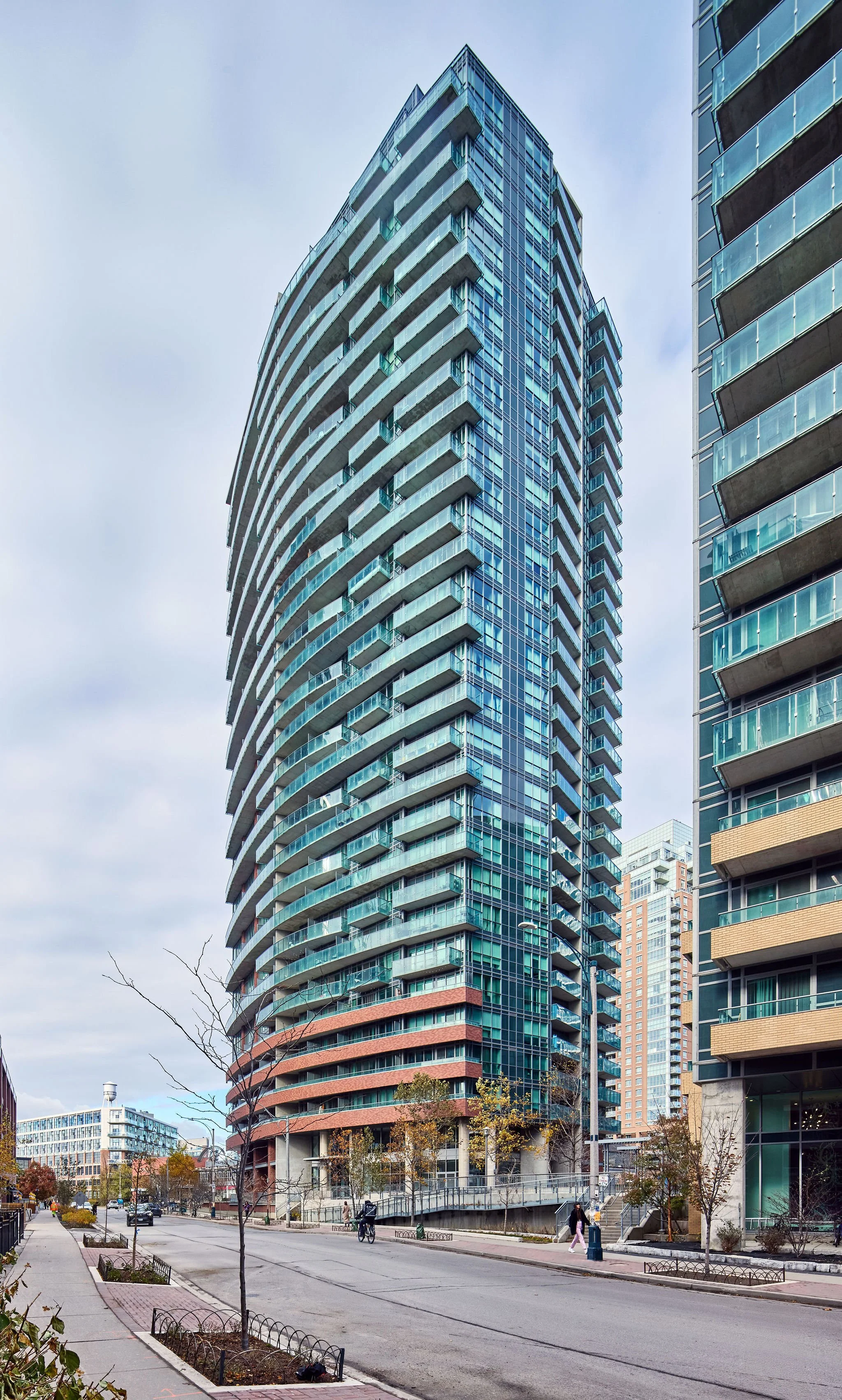A modern high-rise building with curved glass balconies on each floor, set against a cloudy sky. Other tall buildings are visible nearby, with a few people and bicycles on the street below.