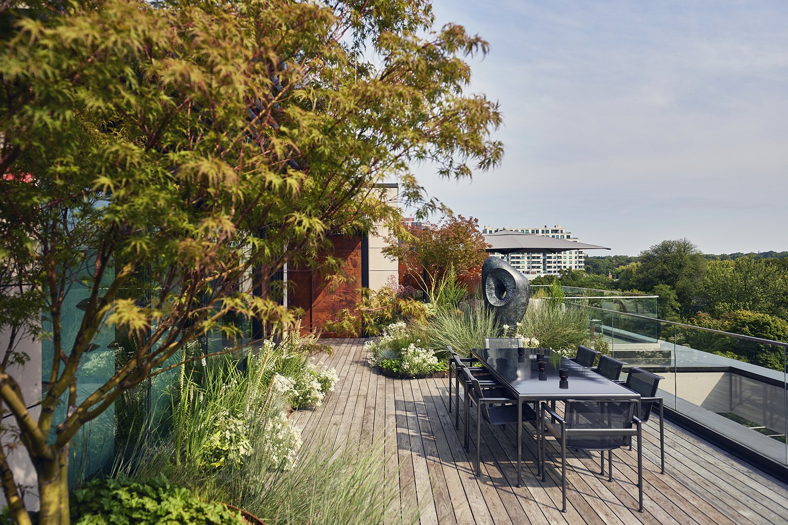 A rooftop terrace with wooden decking, outdoor dining table with chairs, lush container plants, trees, and a modern sculpture. Glass railing offers a view of trees and distant buildings under a clear sky.