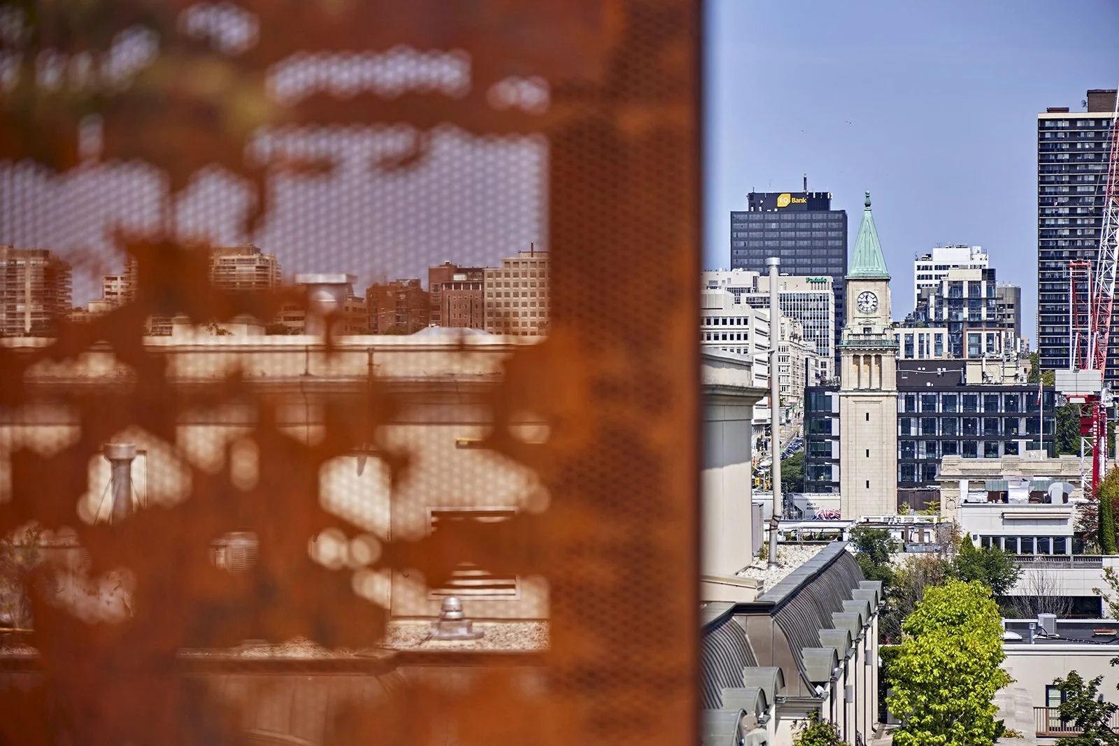 City skyline view with various tall buildings, a clock tower, and a church spire seen through a rusty, perforated metal grate.