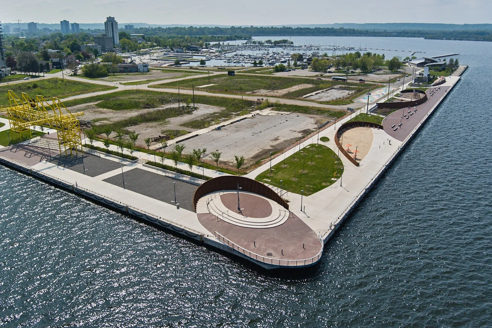 Aerial view of a waterfront park with skateboarding ramps, green areas, walking paths, and a construction site, next to a marina with numerous boats, and a city skyline in the background.