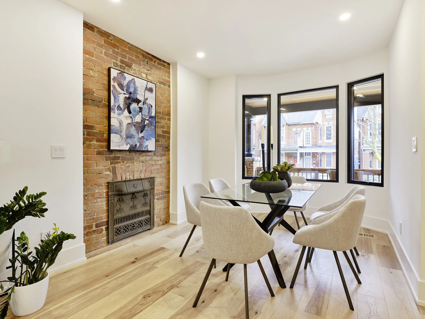 Modern dining room with a glass table and six beige chairs, a brick fireplace with artwork above, large windows looking out to a residential street, wood flooring, and a potted plant in the corner.