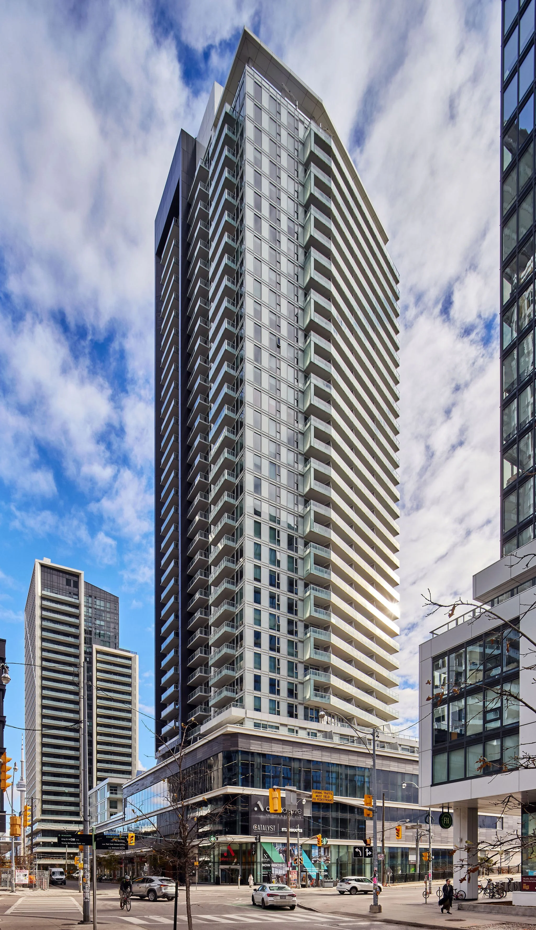 A tall modern high-rise building with glass windows and a combination of protruding and recessed balconies. It is situated on a city street with cars, bicycles, and pedestrians, and other tall buildings can be seen nearby under a partly cloudy sky.
