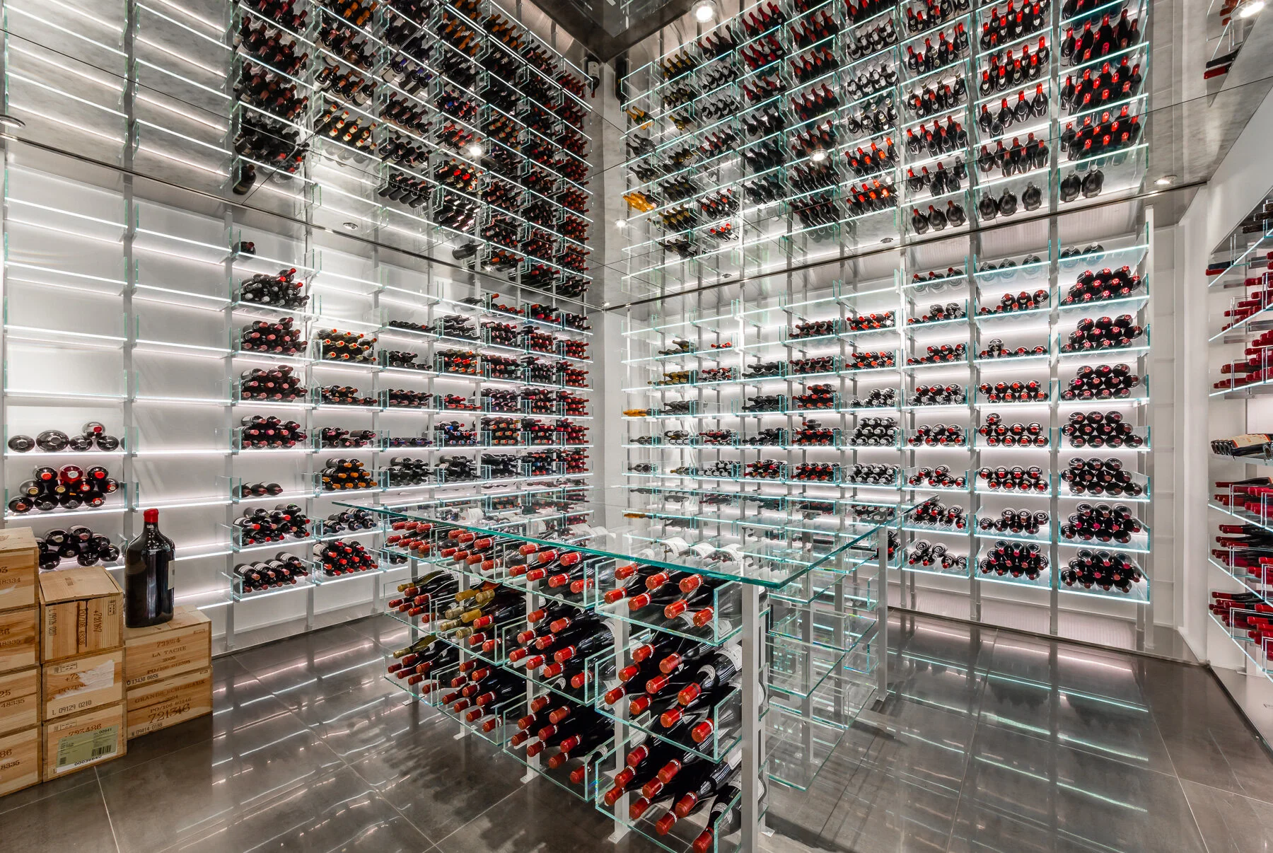 A modern wine cellar with glass and metal shelving filled with bottles of wine, illuminated by built-in lighting, with some cardboard boxes and a large wine bottle on the floor to the left.