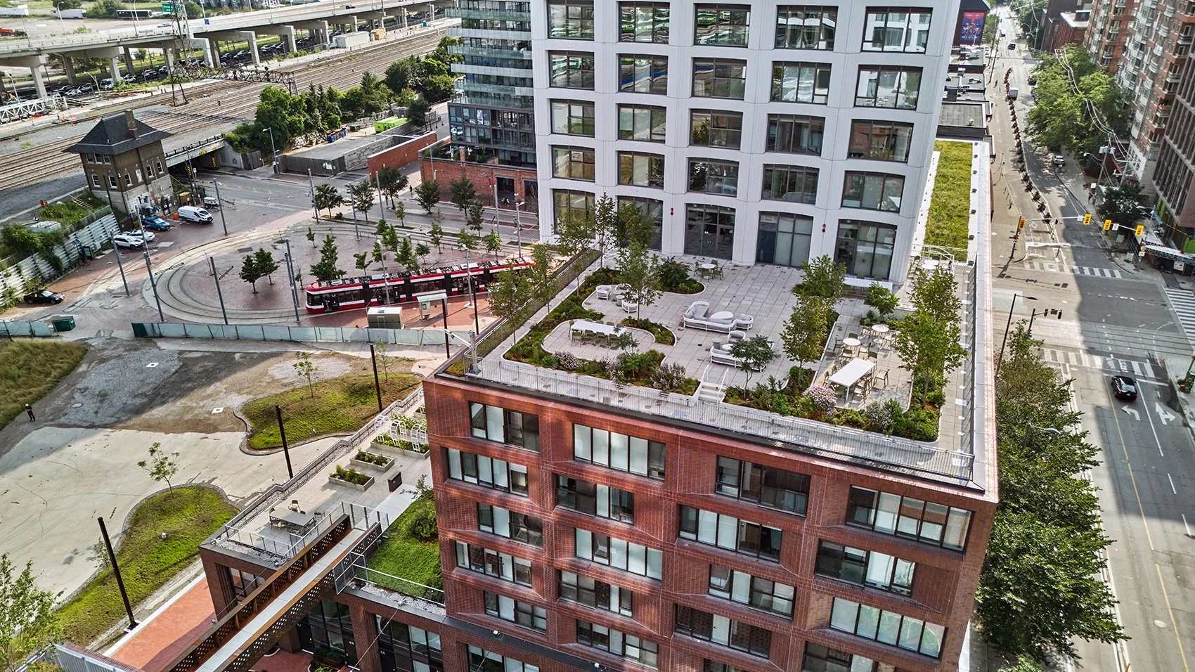 Aerial view of a modern city building with a rooftop terrace landscaped with plants, trees, and outdoor furniture, adjacent to a street with cars and crosswalks, and a nearby train station with a red train passing through.