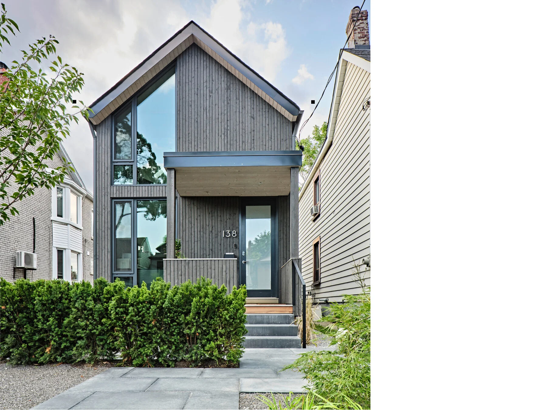 Modern two-story house with dark wood exterior, large front window, and a small front porch with steps and metal railing, located between older houses.