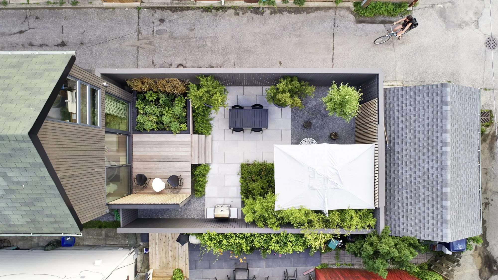 Aerial view of a backyard patio with seating, a white umbrella, surrounded by greenery, with a person riding a bicycle on the street outside the fence.