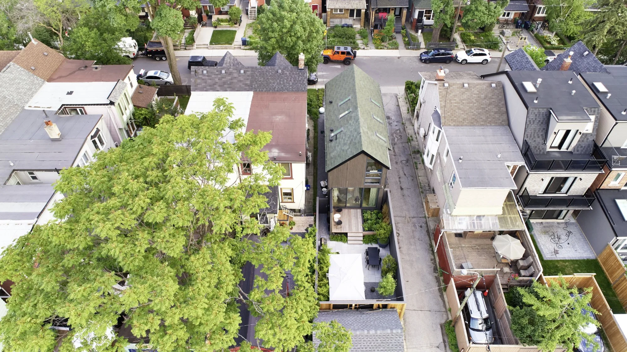 An aerial view of a neighborhood street with houses, trees, and parked cars, including a distinctive small house with a green roof and a backyard patio.