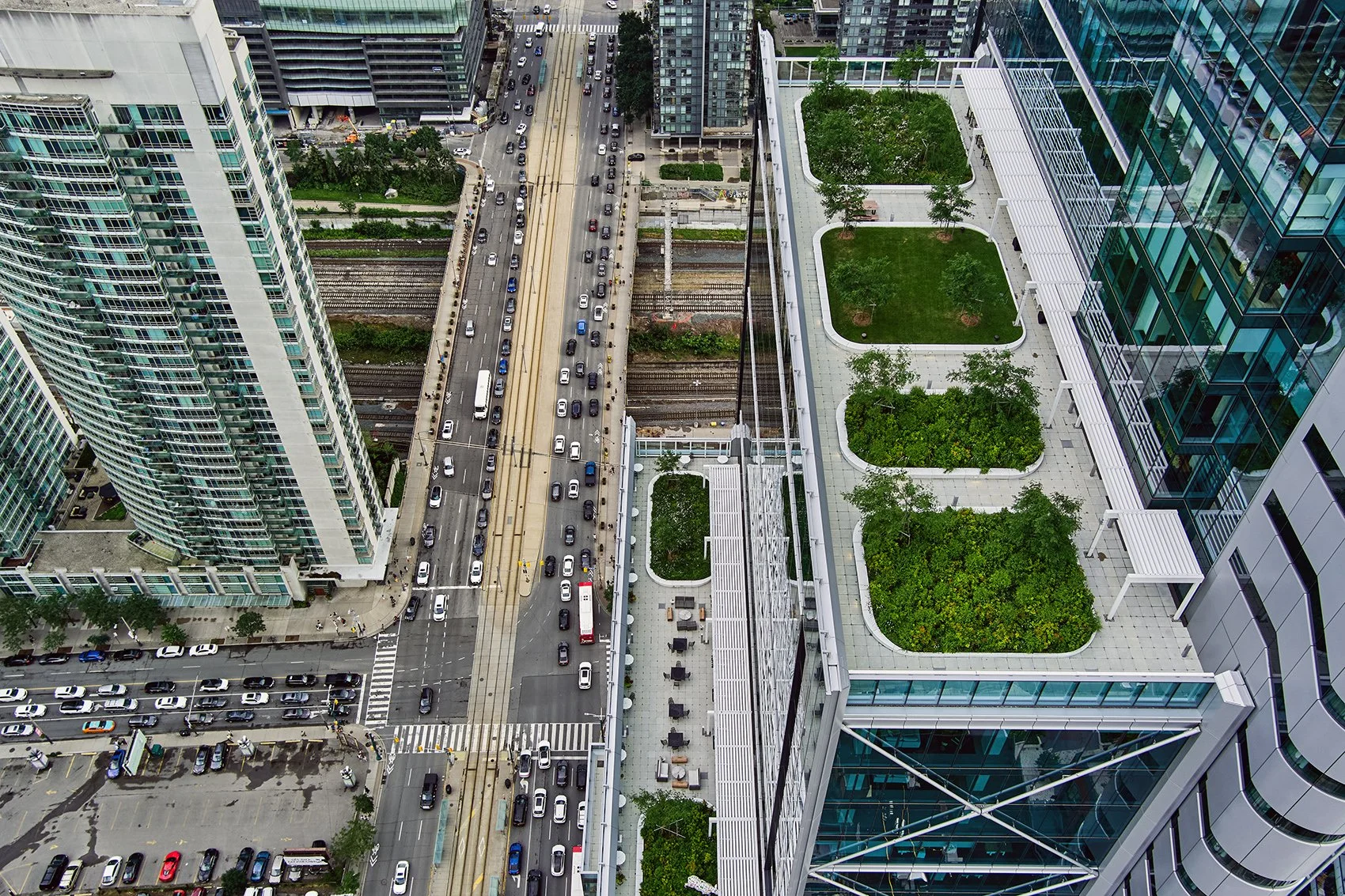 A high-angle view of a city street with multiple cars, a bus, and train tracks, surrounded by tall modern glass skyscrapers and green rooftop gardens.
