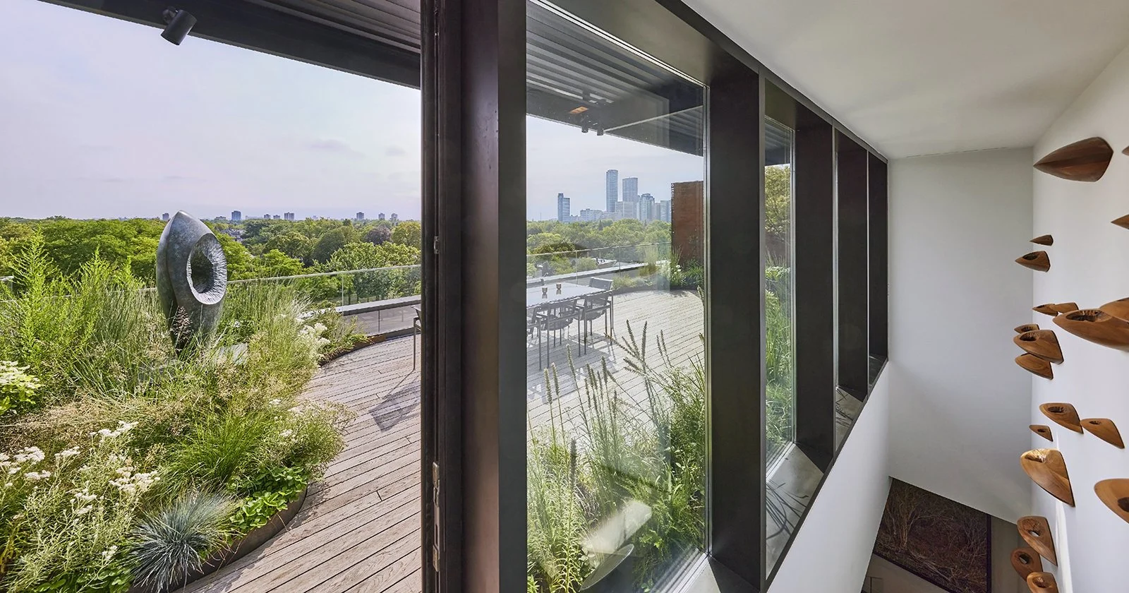 Interior view from a modern home looking out onto a balcony with greenery and city skyline in the distance, featuring glass doors, metallic framing, and a decorative wall art on the right.