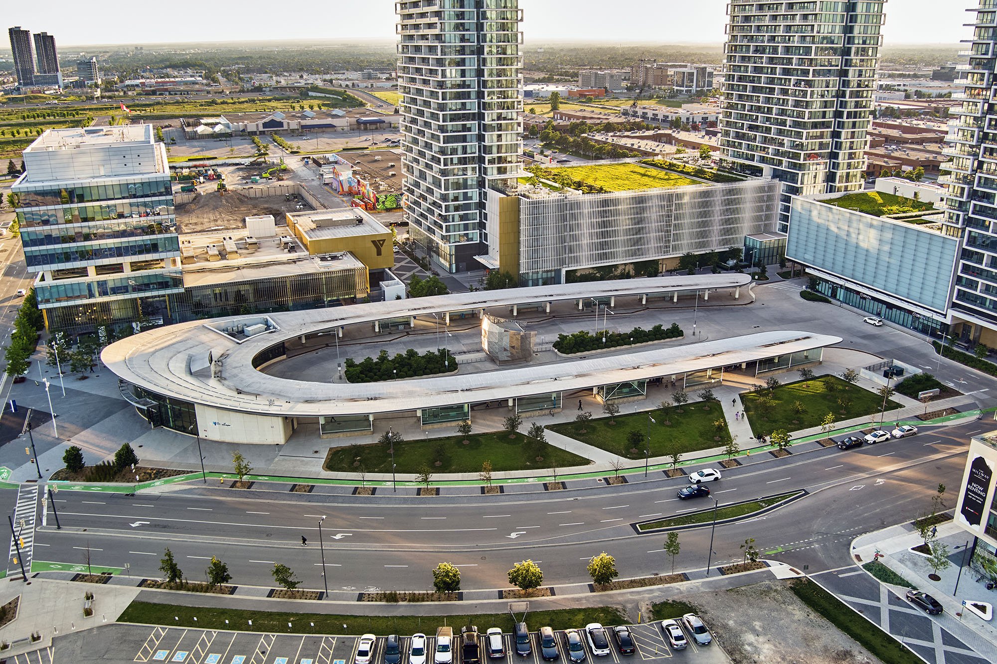 Aerial view of a modern urban area with tall glass buildings, a curved white structure in the foreground, green landscaping, and streets with cars.