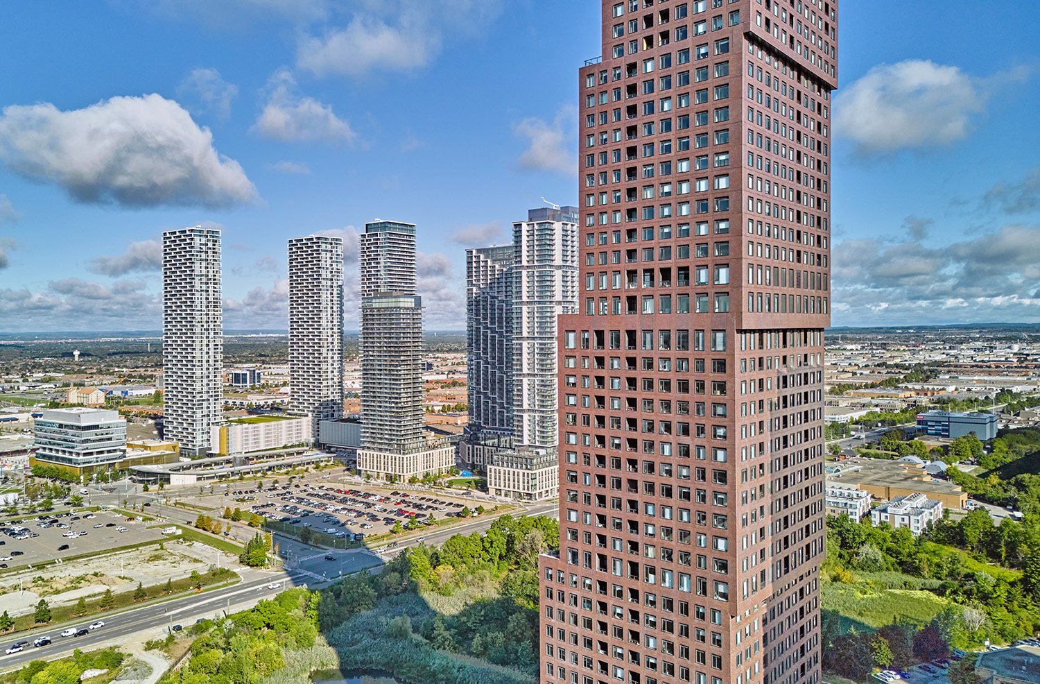 Cityscape with tall residential and office buildings under partly cloudy sky, green trees, roads, and parking lots.