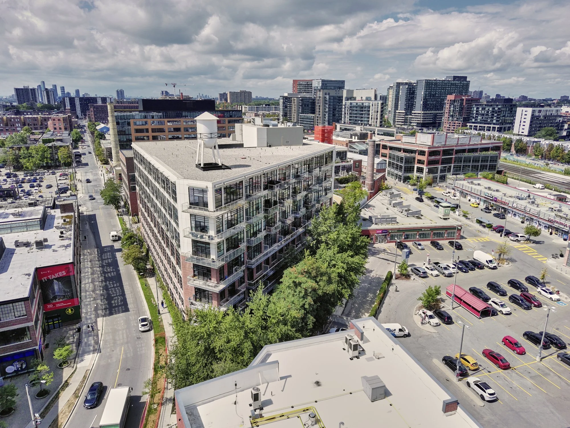 An aerial view of a cityscape showing a multi-story apartment building with balconies, surrounded by roads, parking lots, and various commercial buildings, with a cloudy sky overhead.