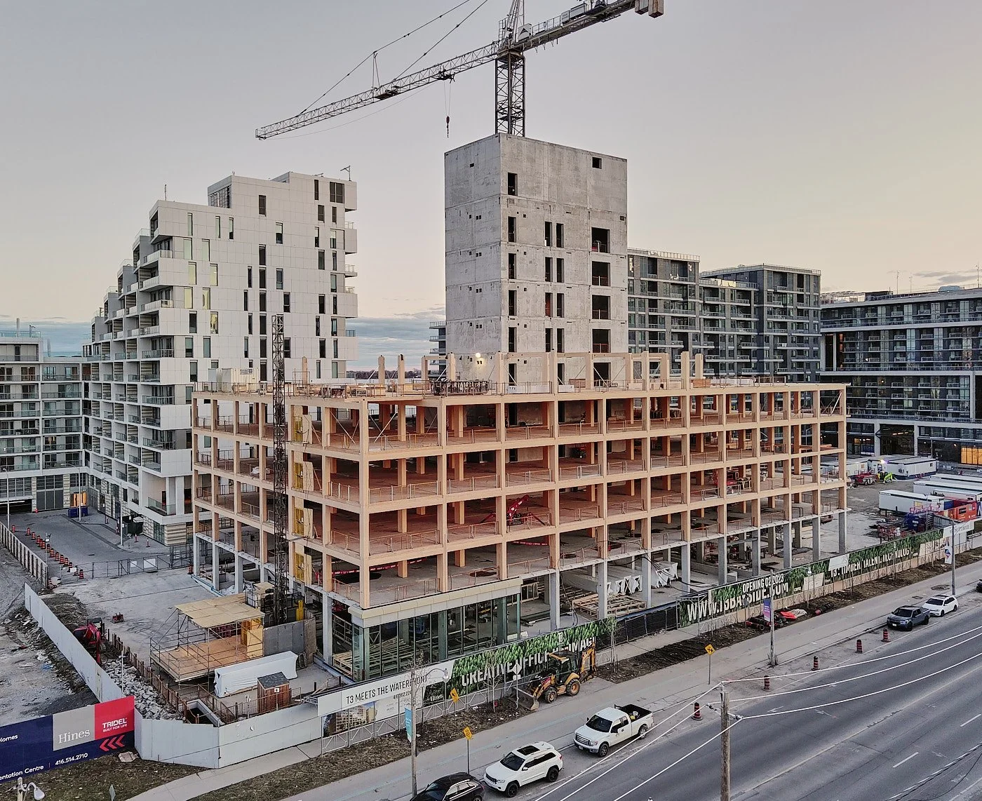 Construction site of a multi-story building with a large crane on top, surrounded by other completed residential buildings, and a road with cars in the foreground.