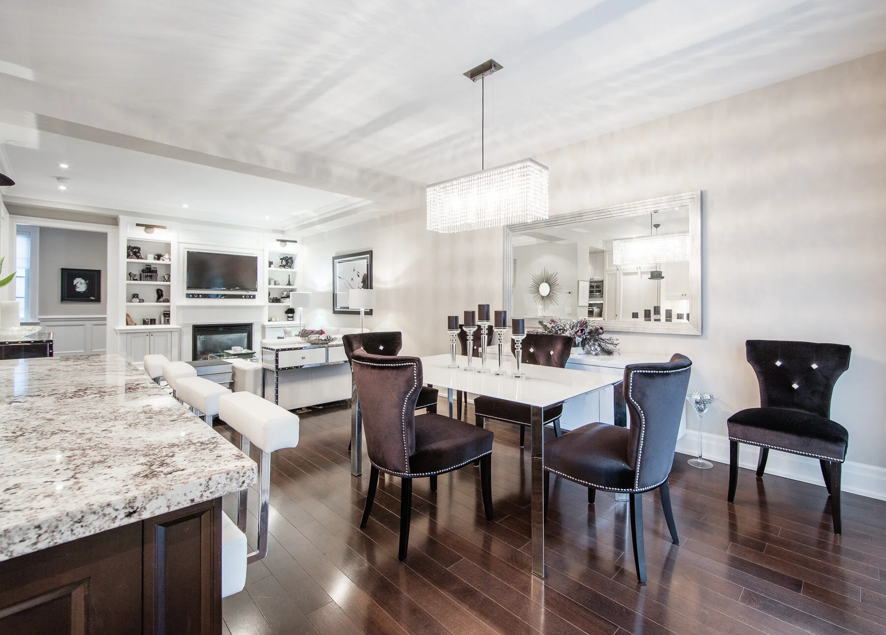 Modern dining area with a white table, six black chairs, and a chandelier, adjacent to a living room with a fireplace, wall-mounted TV, and built-in shelving.