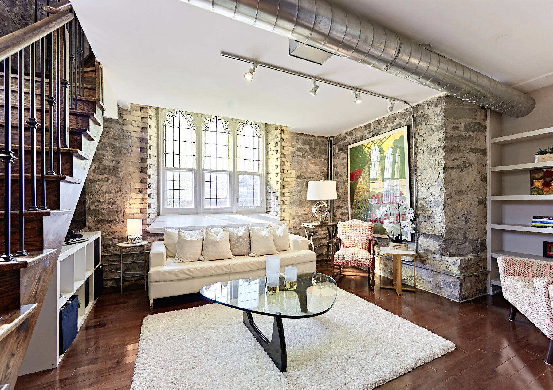 Living room with exposed brick wall, large window, white sofa with beige pillows, glass coffee table, area rug, side tables with lamps, colorful artwork on exposed brick wall, bookshelf with decorative items, and a staircase with wooden steps and bla