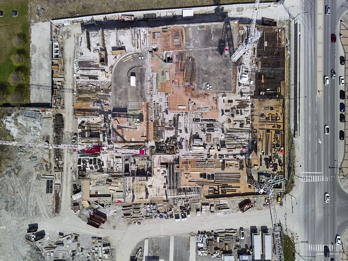 An aerial view of a construction site with building frameworks, cranes, construction materials, and workers, bordered by a road with moving cars.