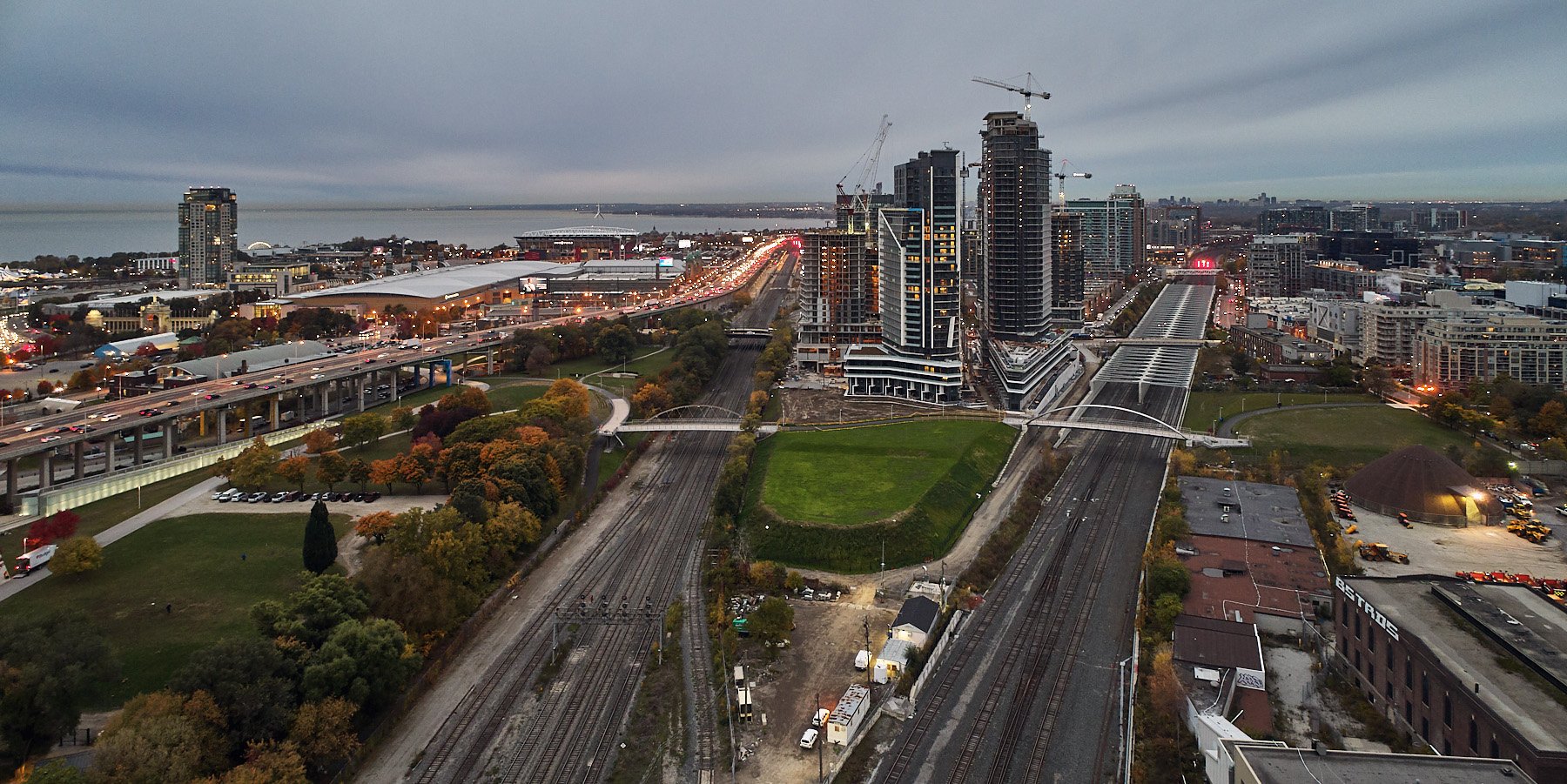 A cityscape at dusk showing high-rise buildings under construction, railroad tracks, a park area with trees, and a highway with traffic.
