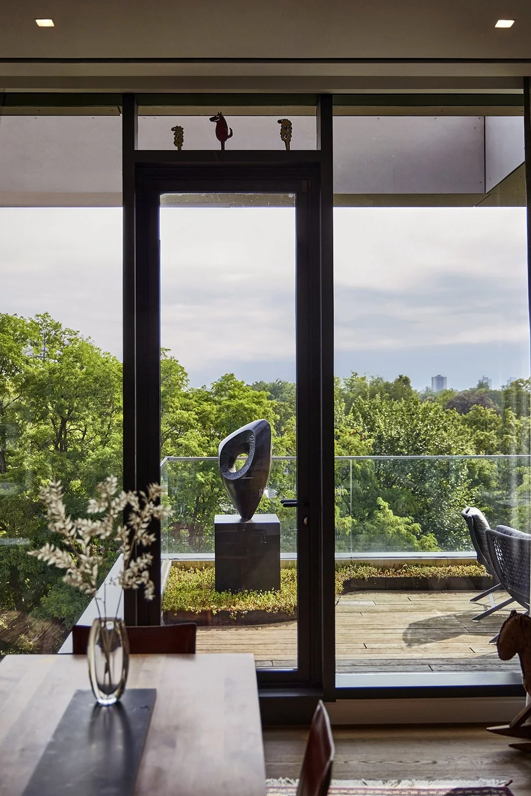 Interior view of a room with a view of a balcony and green trees outside. Inside, there is a wooden dining table with a vase of white flowers, and on the balcony, there is a modern sculpture on a pedestal. Above the door, there are three decorative o