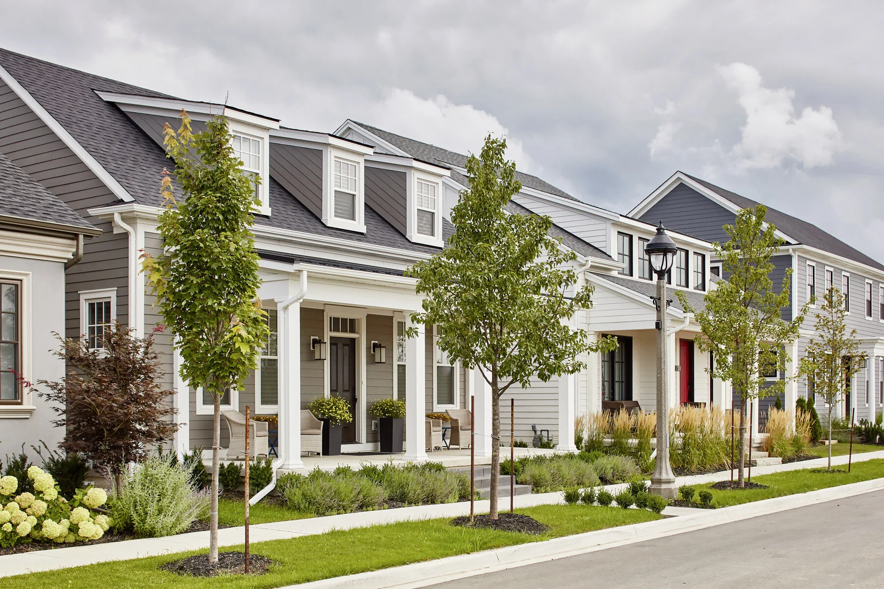 Modern row of townhouses with small front porches, landscaped gardens, young trees, and a street lamp, under cloudy sky.
