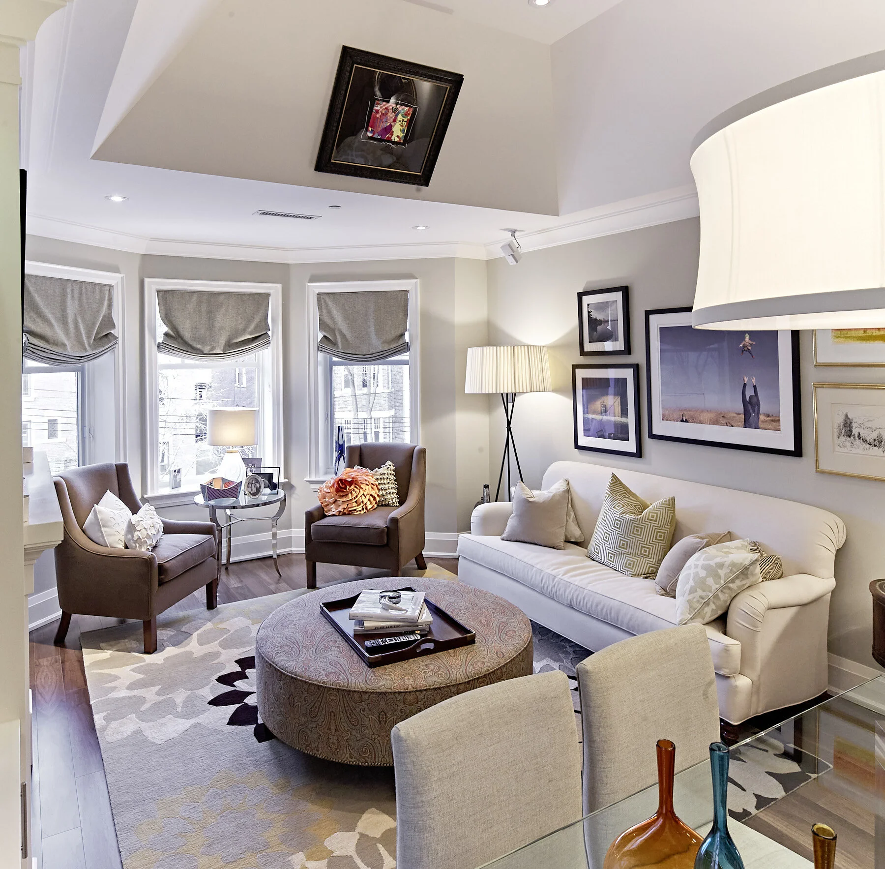 A living room with a white couch, two brown armchairs, a round ottoman, a floor lamp, and framed pictures on the wall. Three windows with beige shades let in natural light.