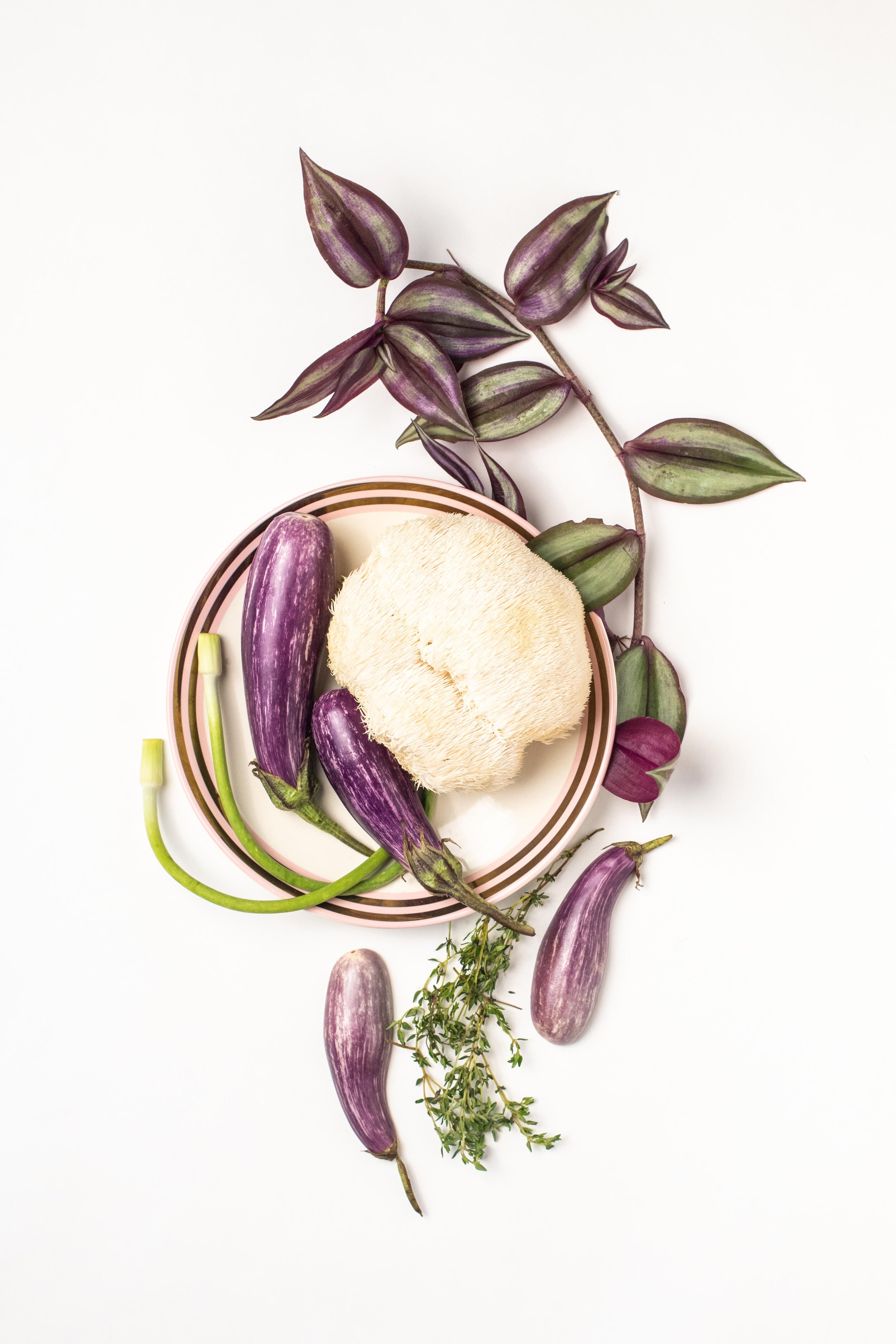 Assorted fresh produce including mini purple eggplants, a lion's mane mushroom, green garlic scapes, sprigs of thyme, and striped purple leaves, arranged on a white surface with a plate.