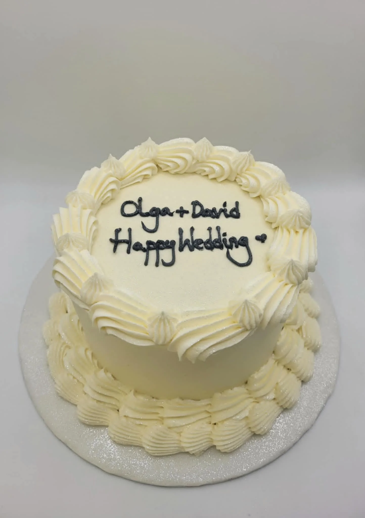 White wedding cake with piped cream decorations, inscribed with 'Olga + David Happy Wedding' in black icing.
