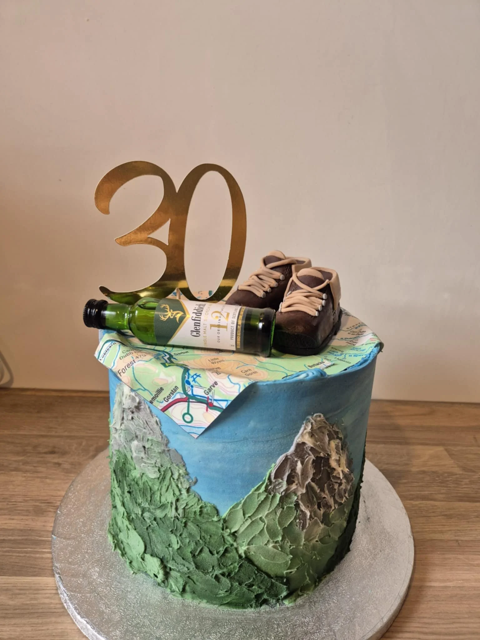 Celebration cake with mountain and forest scenery, topped with a large gold '30' cutout, a map, a small bottle of Glenfiddich whiskey, and a pair of small shoes.