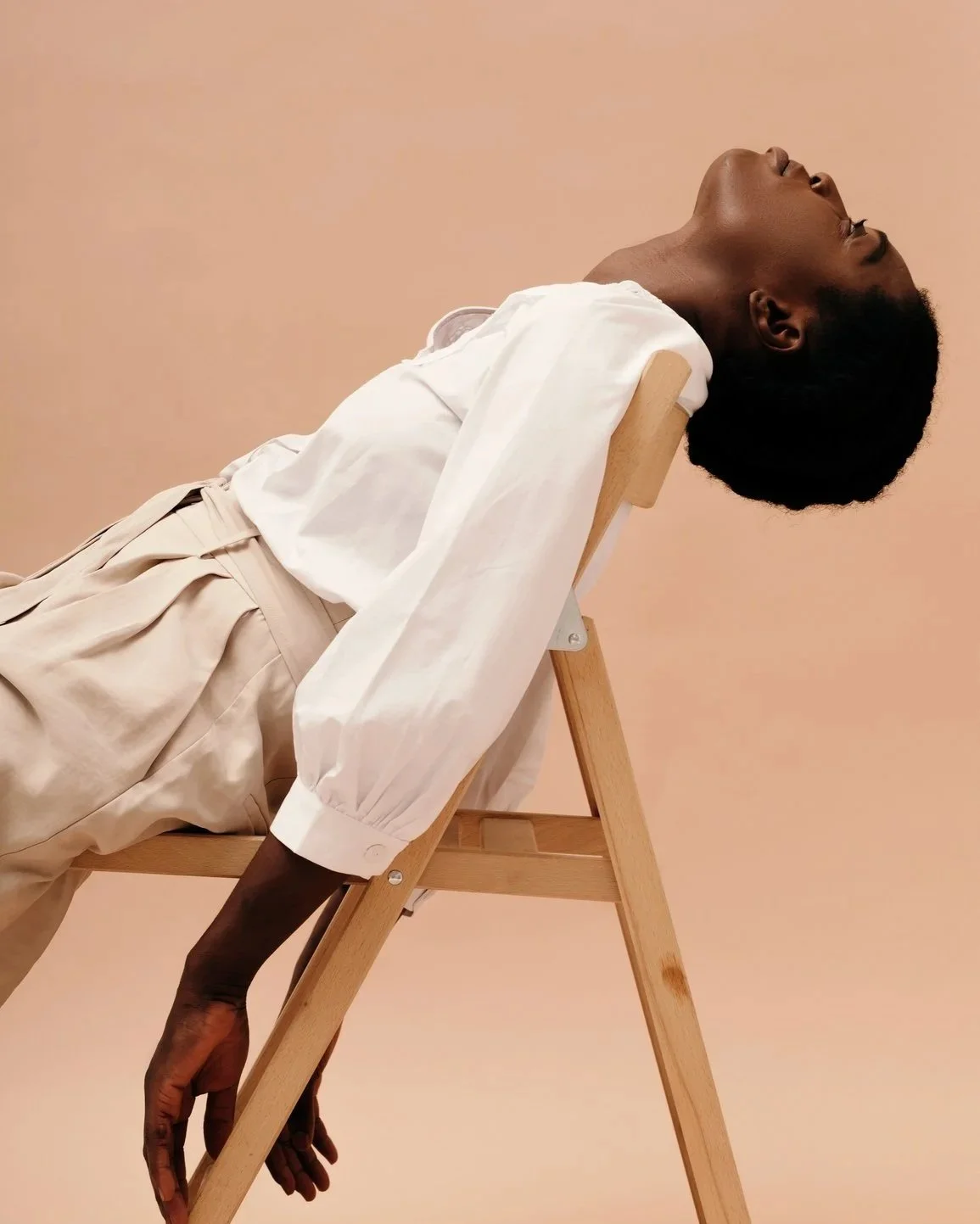 A person with dark skin and short curly hair is leaning back on a wooden chair with their eyes closed, head tilted upward, against a plain peach-colored background, wearing a white shirt and beige pants.