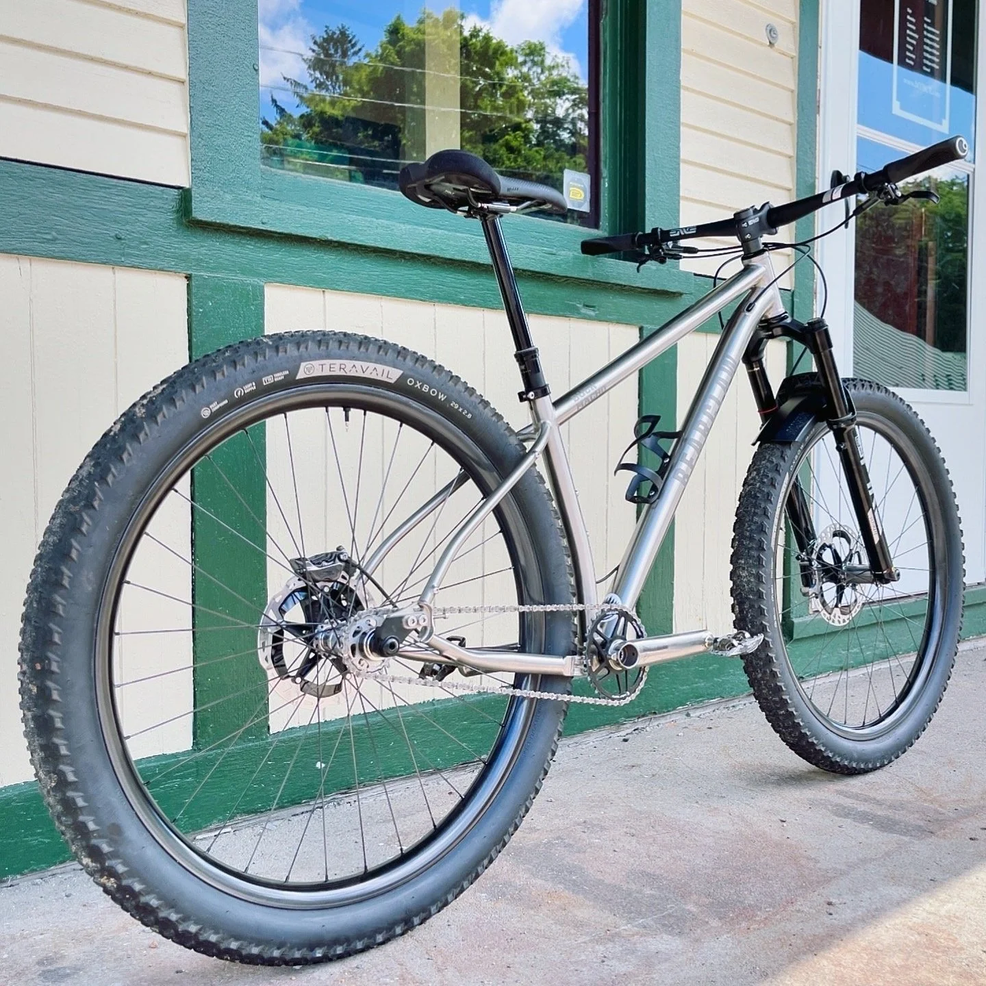 A silver mountain bike with knobby tires, parked outside a building with green and white trim and glass windows.