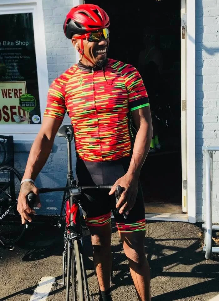 Smiling man in cycling gear, red helmet, sunglasses, holding a road bike, standing outside a bike shop.