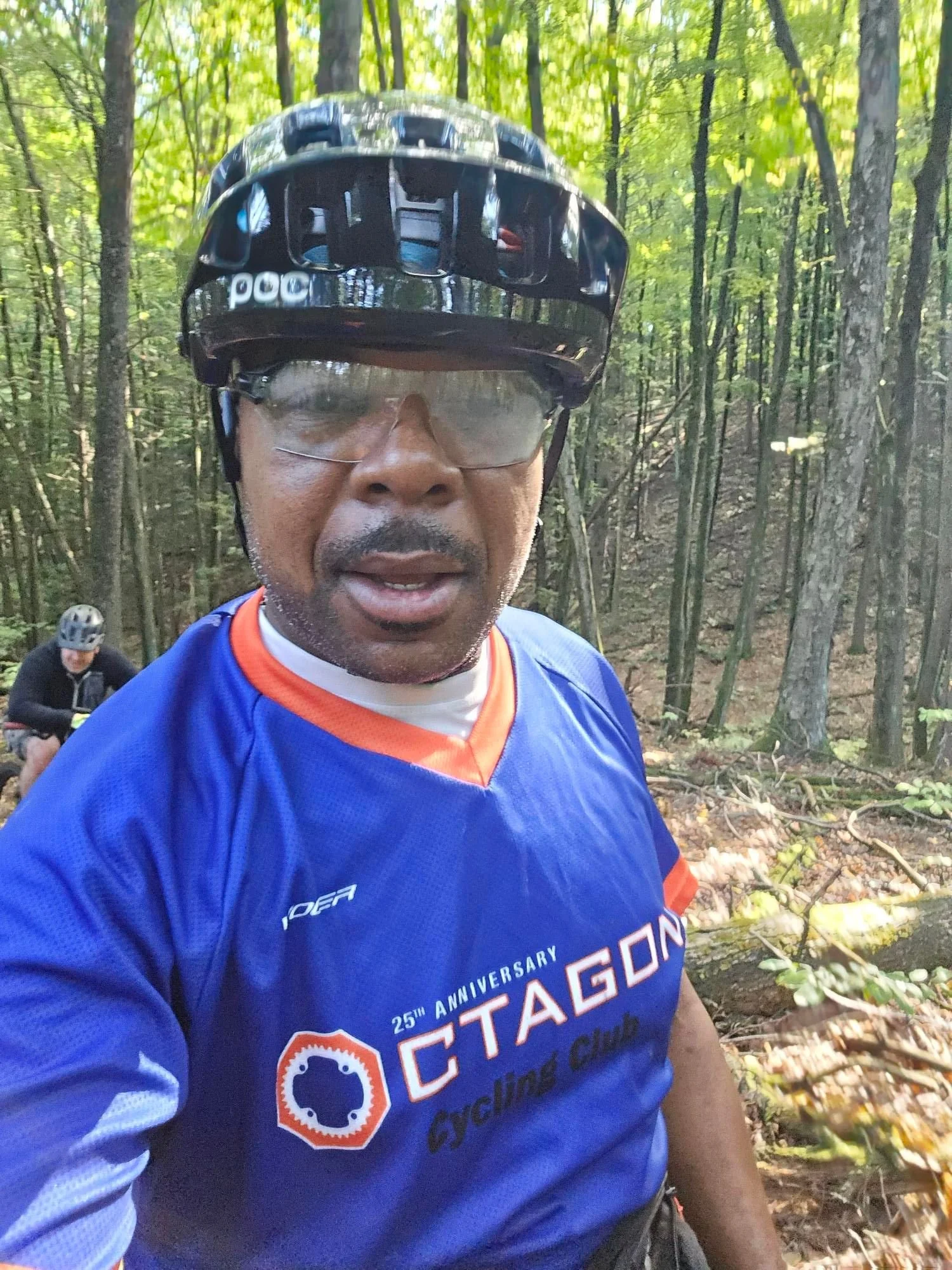 A man wearing a black helmet and protective glasses taking a selfie in a forest. He is dressed in a blue cycling jersey with orange accents and a logo that says 'CITAGON Cycling Club'. A person in the background is also in cycling gear, sitting on the ground among trees.