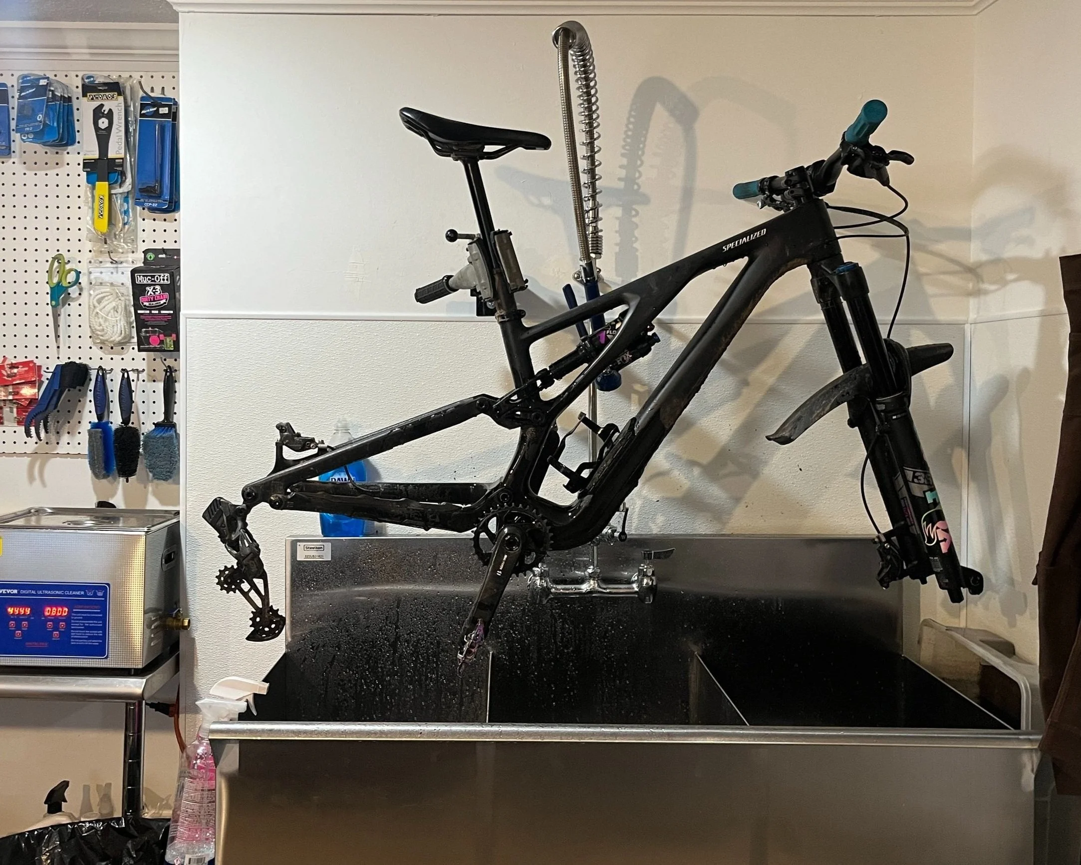 A black mountain bike is being washed in a stainless steel sink in a garage or workshop, with the bike mounted on the sink's edge and water running over it.