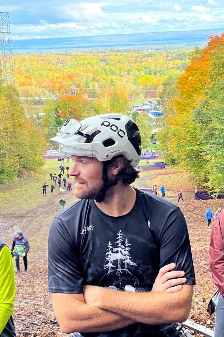 A man wearing a black t-shirt and a white helmet standing outdoors, with people and colorful fall trees in the background.