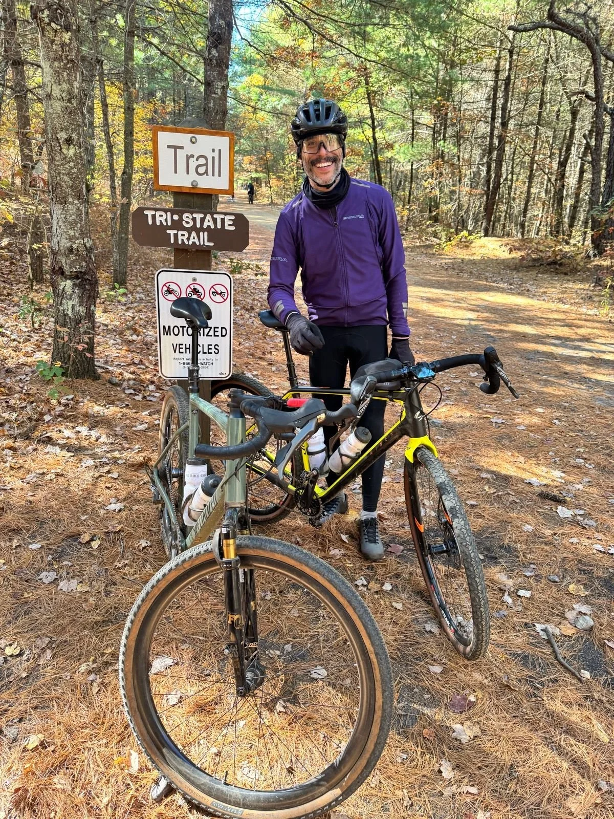 A man in biking gear, including a purple jacket and helmet, standing next to his mountain bike on a dirt trail in a wooded area during autumn. There are trail signs, including one that says 'Trail' and another 'Tri-State Trail,' and a sign indicating motorized vehicles are not allowed.