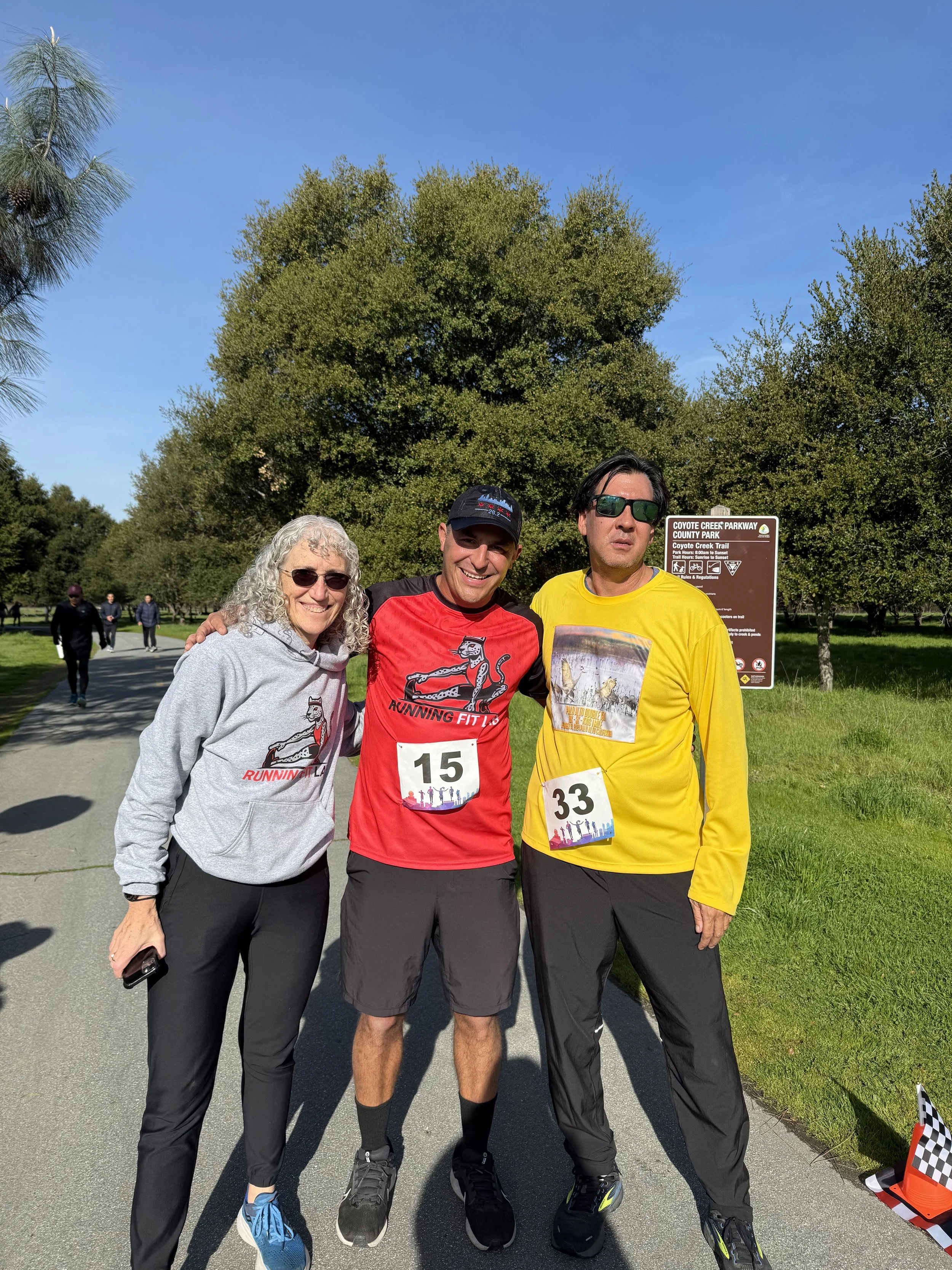 Three people posing together outdoors on a pathway in a park, wearing running attire with numbered bibs. Trees and a park sign are visible in the background.