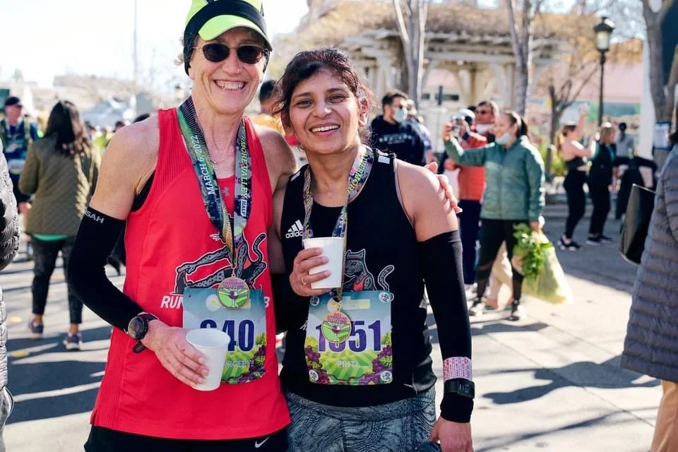 Two Running Fit Lab runners wearing medals and race bibs smiling while holding cups, after finishing Livermore Half Marathon