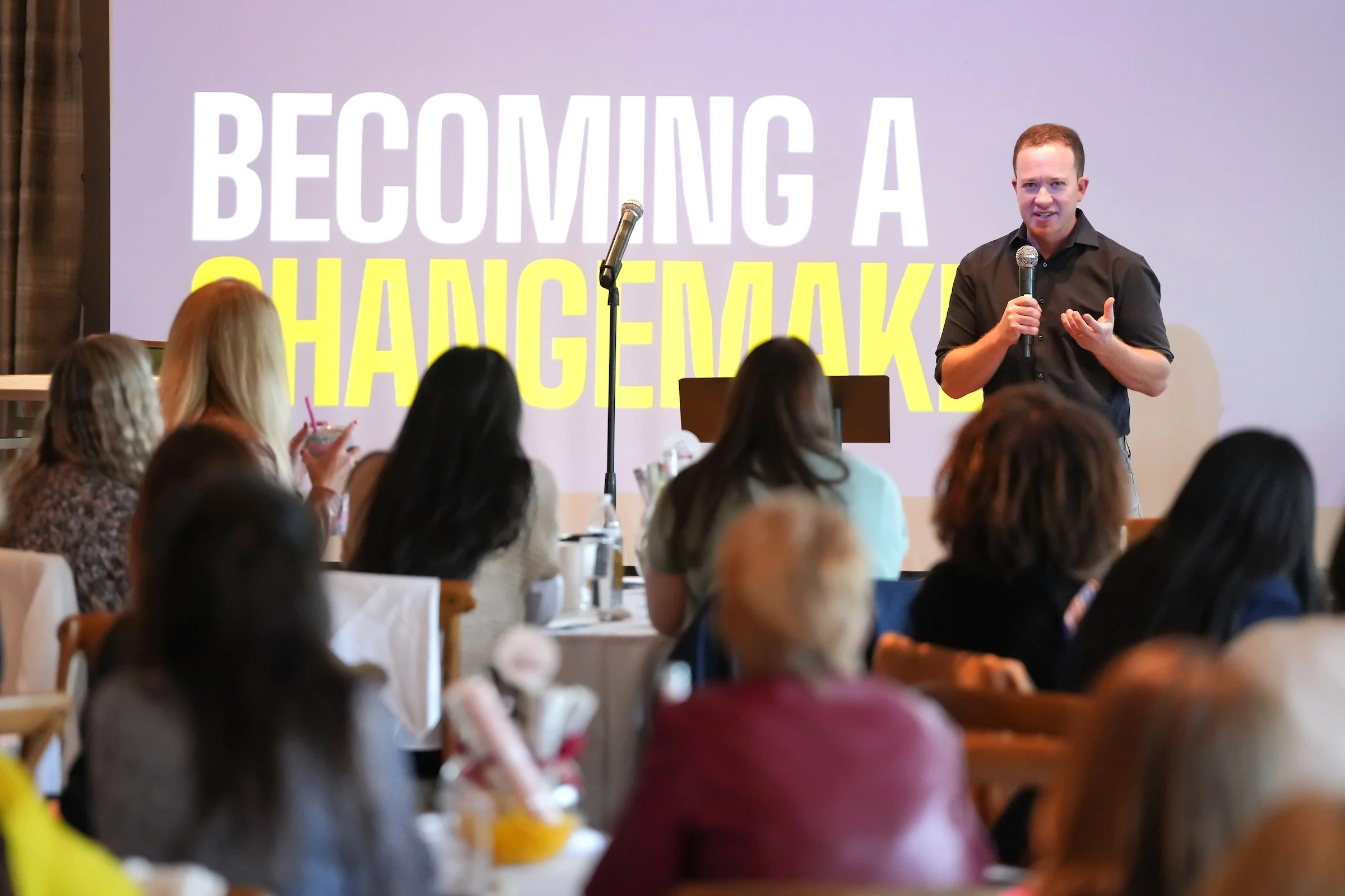 Man giving a speech at a conference titled 'Becoming a Changemaker' with an audience of diverse women seated at tables.