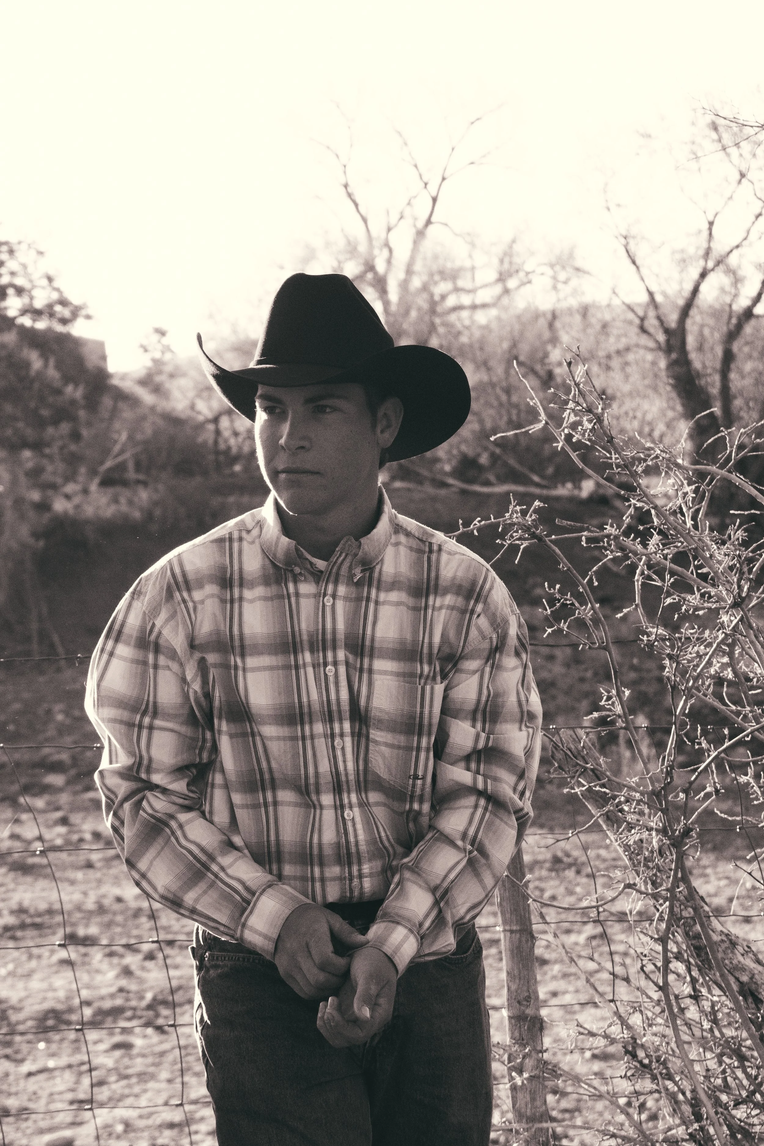 Young man wearing a plaid shirt and cowboy hat, standing outdoors near a fence and leafless trees, in black and white.