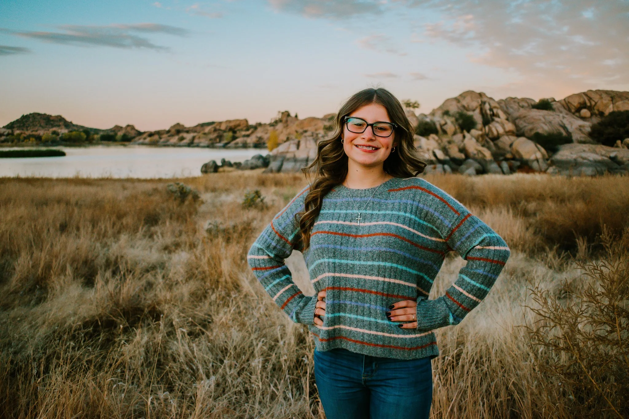 Young woman with glasses and long curly hair standing with hands on hips in a grassy field with a lake and rocky hills in the background during sunset.