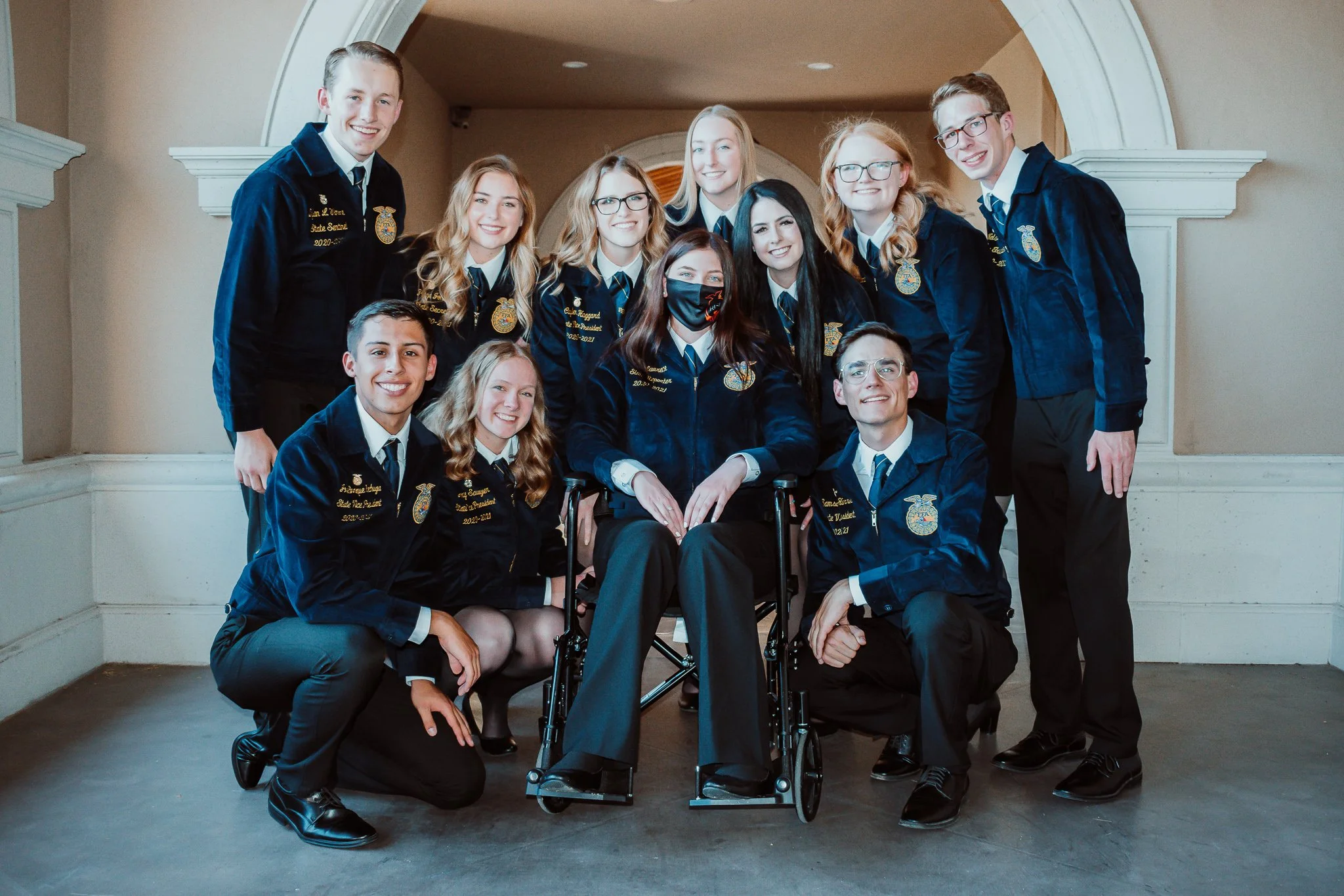 Group of young people in FFA jackets, some kneeling and some standing, posing for a photo indoors.