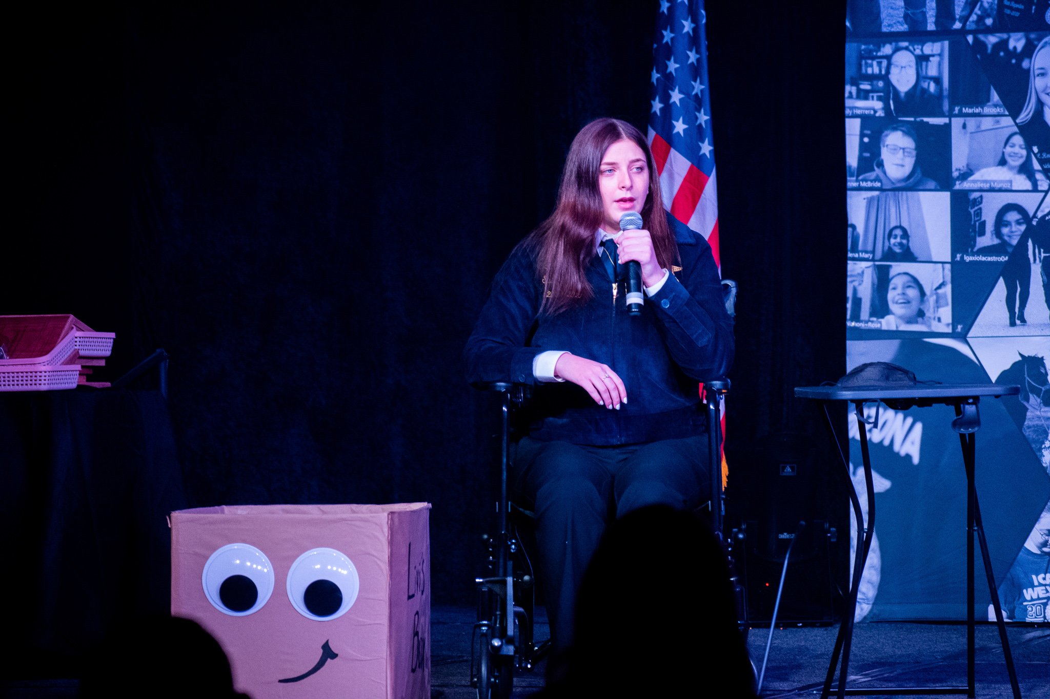A young woman with long brown hair, sitting in a wheelchair, holding a microphone and speaking on stage at an event, with an American flag behind her and a large screen displaying virtual participants to her right.