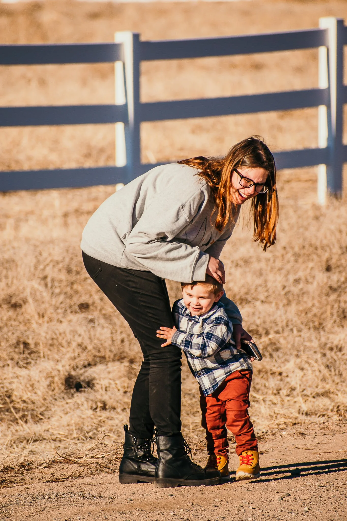 A woman with glasses laughing as she bends over, hugging a young boy with light brown hair and a plaid shirt, outside on a dirt path with a wooden fence in the background.