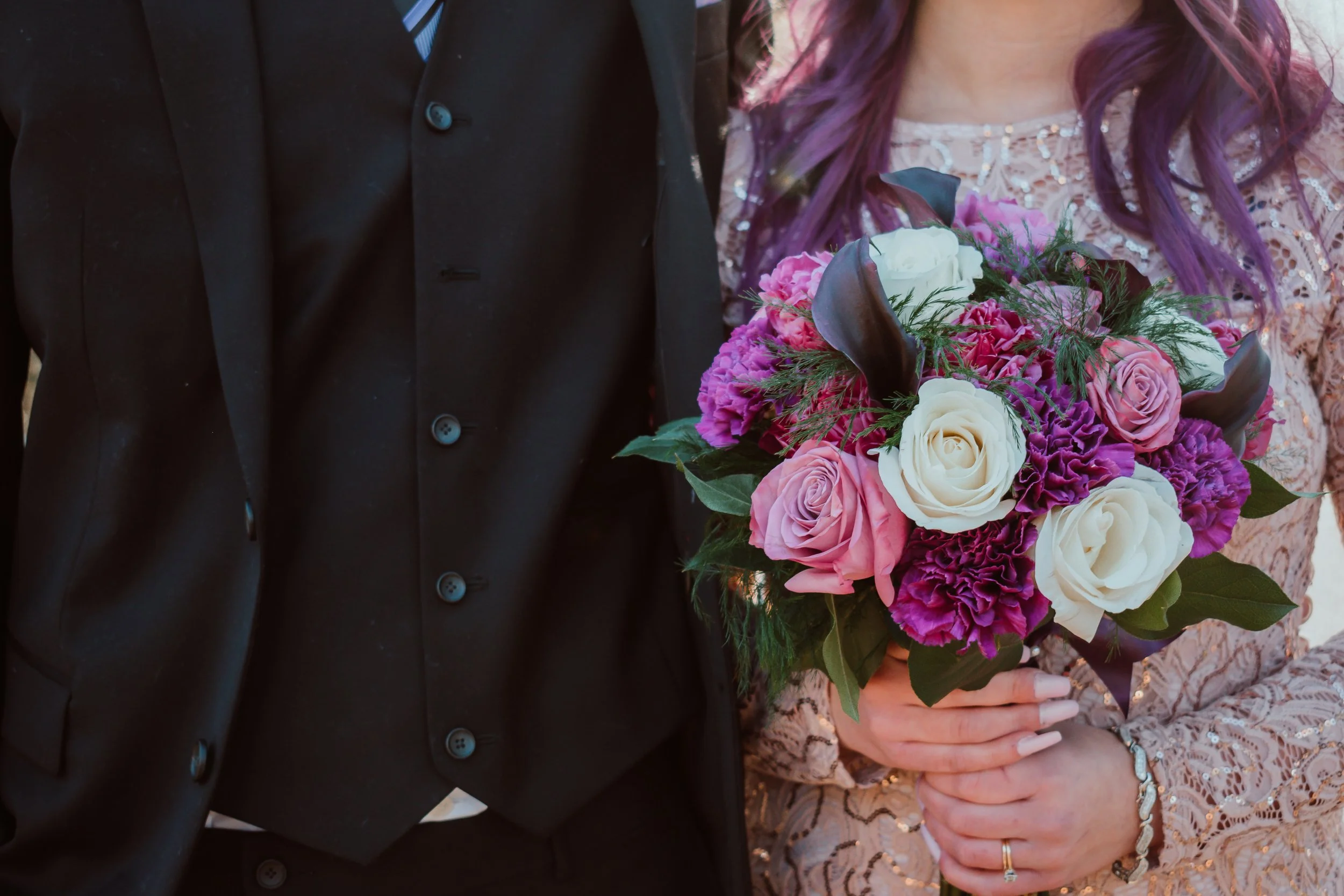 A bride holding a bouquet of pink, white, and purple flowers, standing next to a groom dressed in a black suit.
