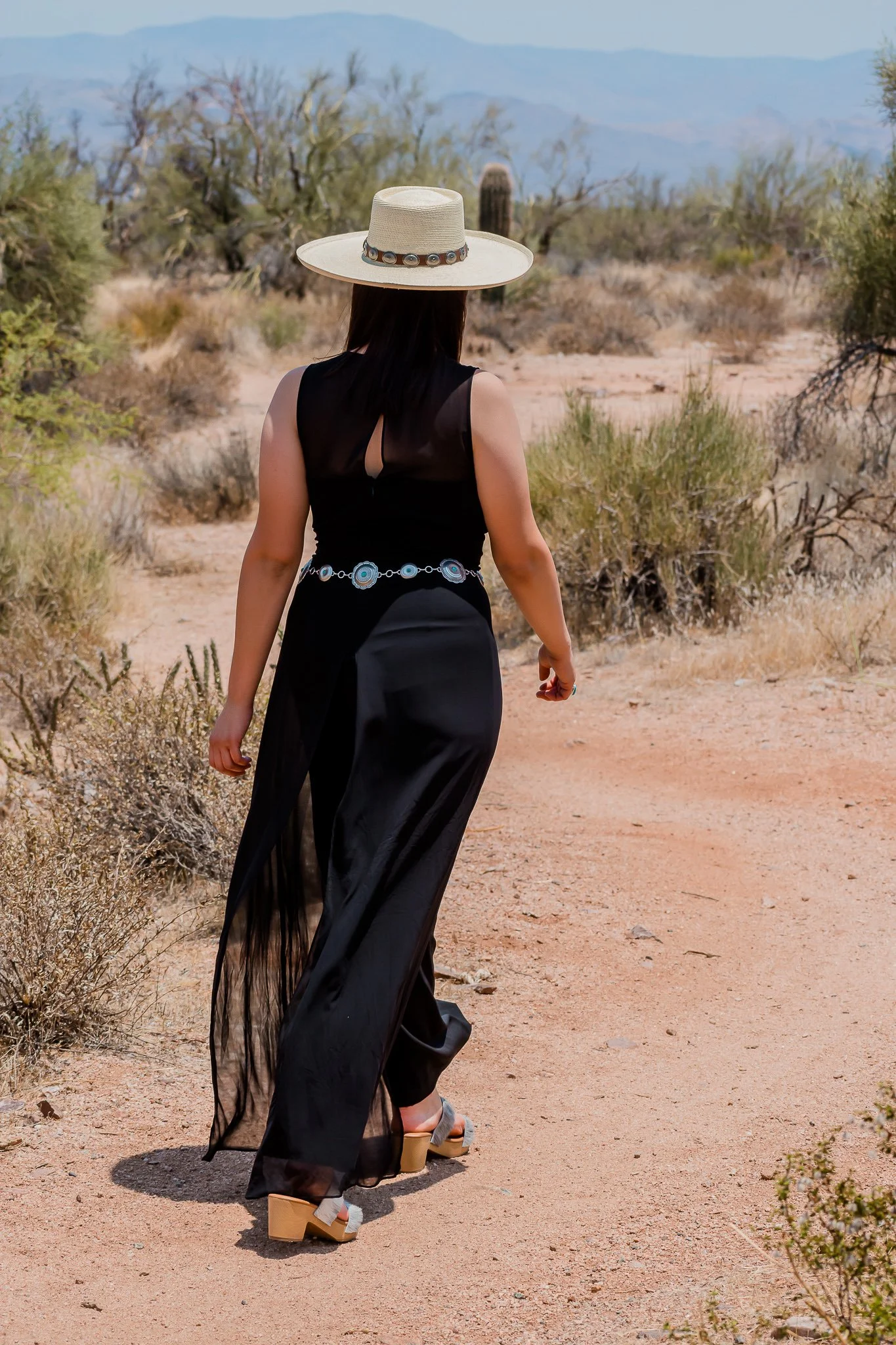 A woman walking through a desert landscape wearing a wide-brimmed hat, a black flowing dress, and high-heeled shoes.