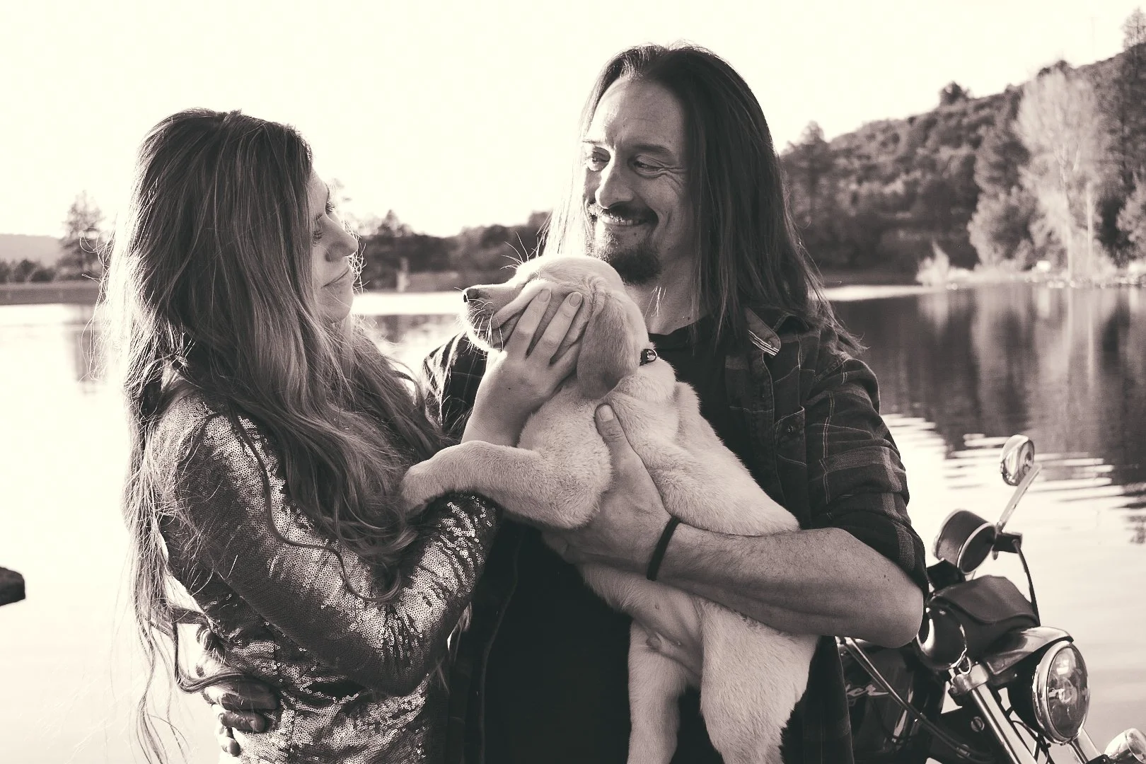 A young woman and a man holding a puppy by a lake during sunset.