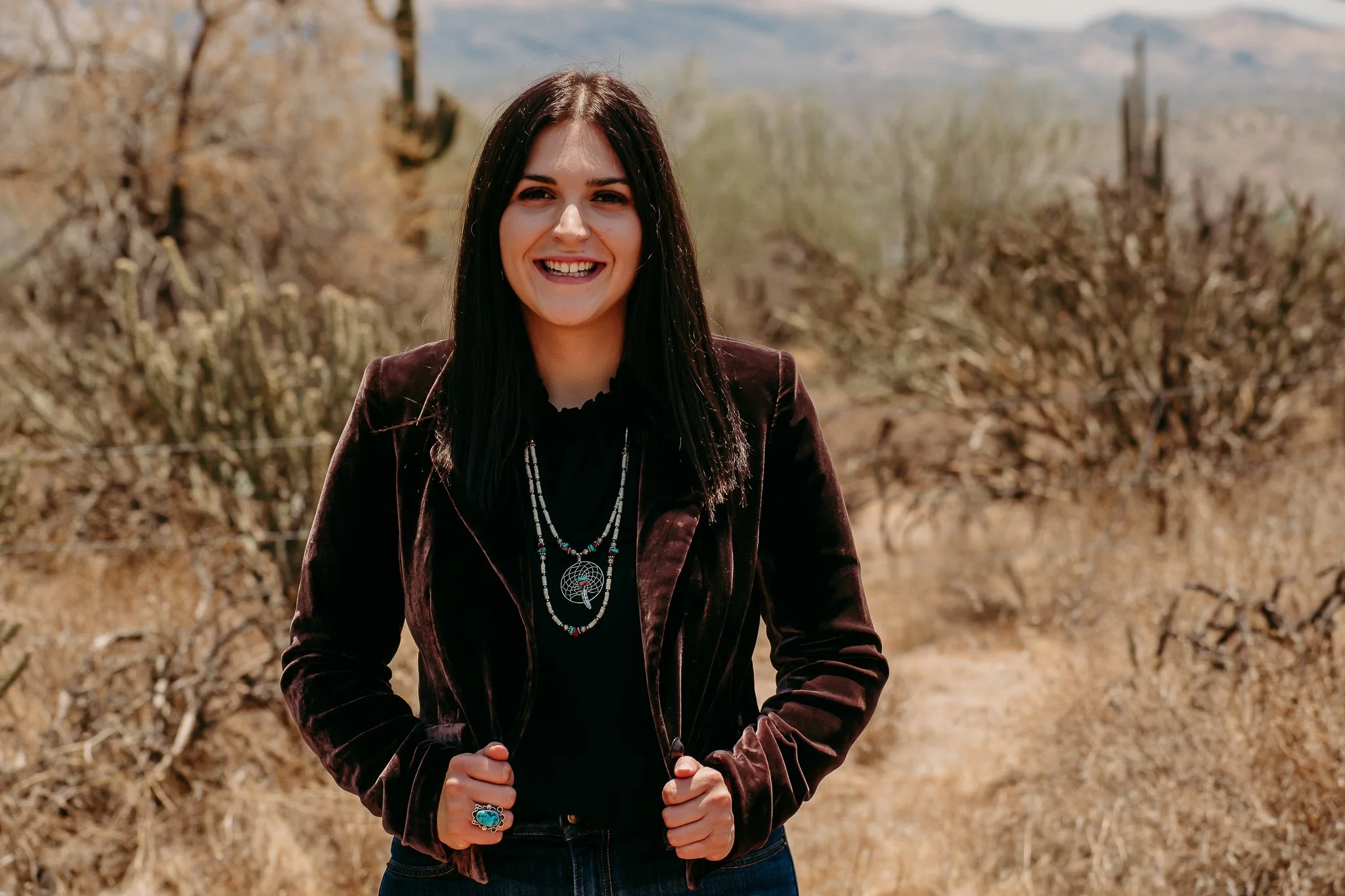 A woman with dark hair smiling outdoors in a desert landscape with cacti and bushes.
