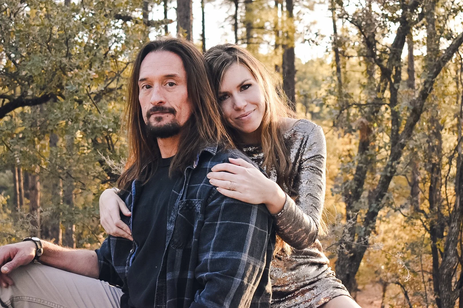 A man and woman sitting outdoors in a forest during autumn, with the woman resting her hand on the man's shoulder. The man has long hair, a beard, and wears a black shirt and plaid jacket. The woman has long hair and wears a shiny, patterned dress, s