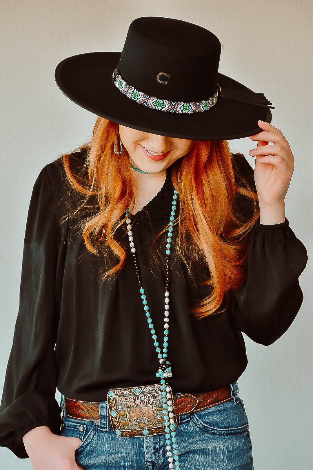 A woman with long red hair wearing a black hat, black blouse, blue jeans, and a large belt, smiling with her face mostly hidden by the hat.