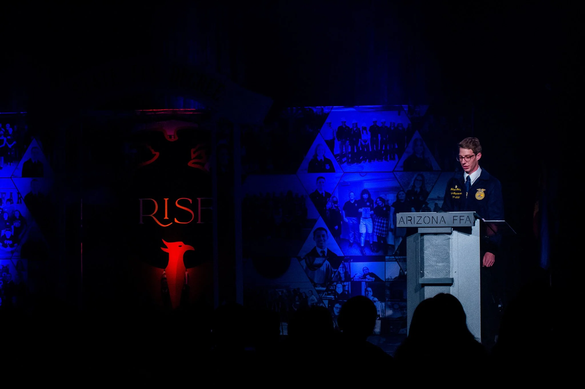 A young man in a dark suit and glasses speaking at a podium with a sign reading 'ARIZONA FFA' during a ceremony or event, with photo collages and a large banner with the word 'RISE' and a phoenix symbol in the background.