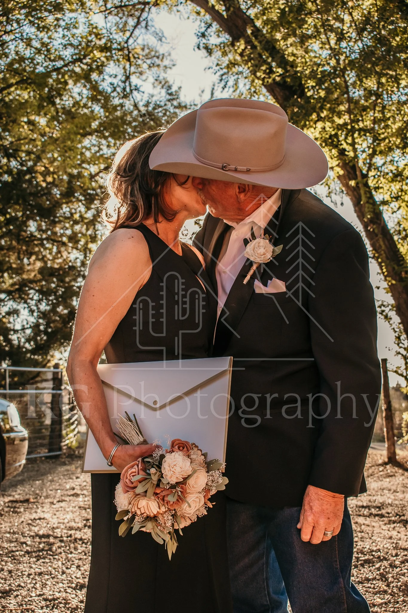 A couple, dressed in formal attire, sharing a kiss outdoors during sunset. The woman is holding a bouquet and a framed item, and the man is wearing a large cowboy hat. Trees and sunlight create a warm background.